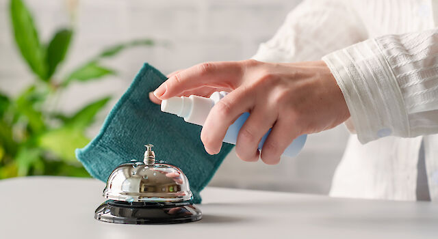 Person holds disinfectant spray and cloth and cleans hotel reception bell with it