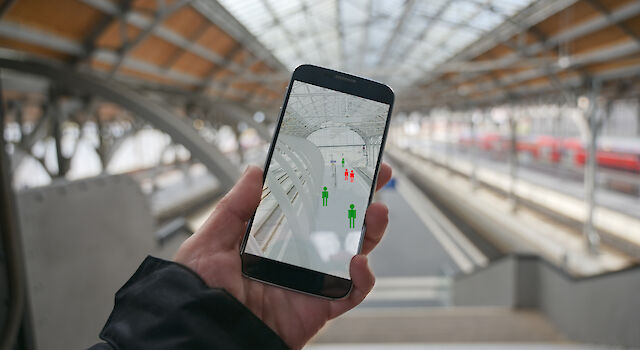 Hand hält Smartphone, auf dem Display sind grün und rot eingefärbte Grafiken von Personen zu sehen. Im Hintergrund verschwommen ein Bahnhof.