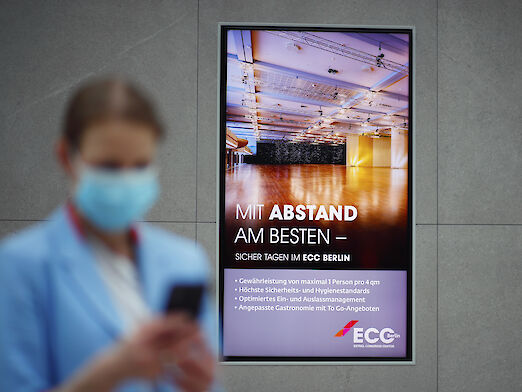 Frau mit Smartphone und Mundschutz vor digitalem Display des ECC Berlin
