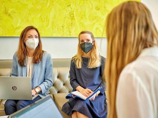 Drei Frauen in Business Outfits, die alle einen Mund-Nasen-Schutz, tragen, sitzen mit ihren Laptops zu einer Besprechung zusammen.