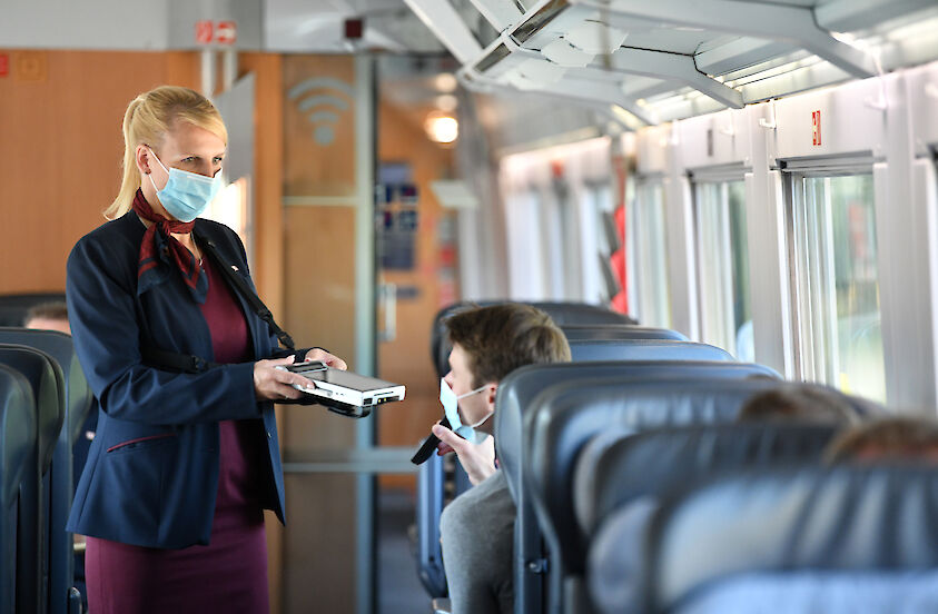 Zugbegleiterin der Deutschen Bahn in Uniform und mit Mund-Nasen-Bedeckung spricht mit einem Fahrgast. | © Deutsche Bahn