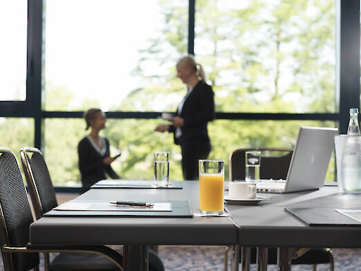 Meetingraum mit Tischen, Laptop, Getränken und zwei Frauen in Business Outfits im Hintergrund.