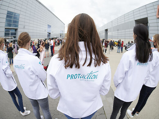 Mitarbeiterinnen auf dem Düsseldorfer Messegelände tragen Jacken mit Aufdruck des Hygienekonzept "PROTaction".