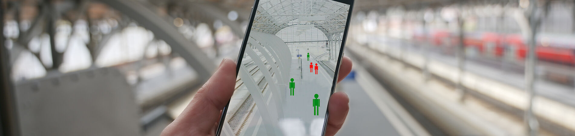 Hand holds smartphone, on the display are green and red colored graphics of people. In the background a train station is blurred.
