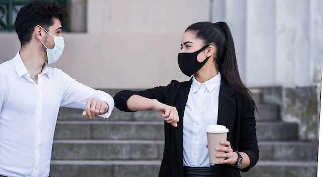 Zwei Geschäftsleute tragen Gesichtsmasken und stoßen zur Begrüßung im Freien die Ellbogen aneinander.
