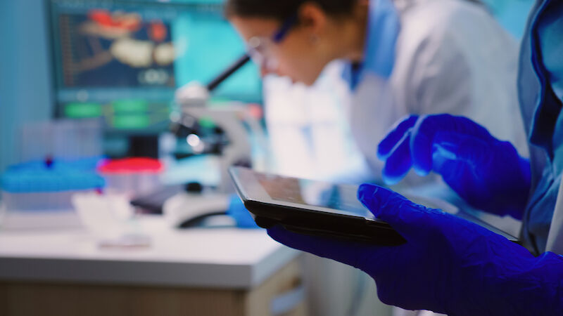 Nahaufnahme einer Person in einem Labor, die Schutzhandschuhe trägt und ein Tablet hält. Eine weitere Person in Laborkleidung im Hintergrund blickt in ein Mikroskop.